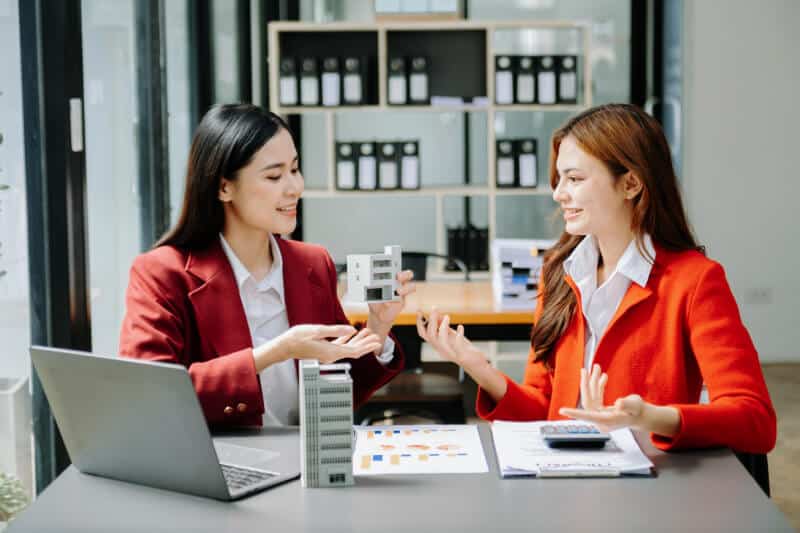 Two women in red blazers discuss real estate at a desk with a laptop, model buildings, and documents in a modern office setting.
