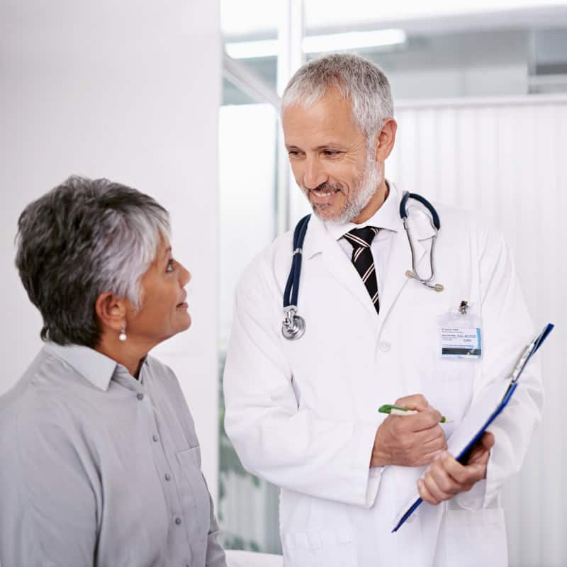 A doctor in a white coat holding a clipboard talks to an older woman patient in a medical office.