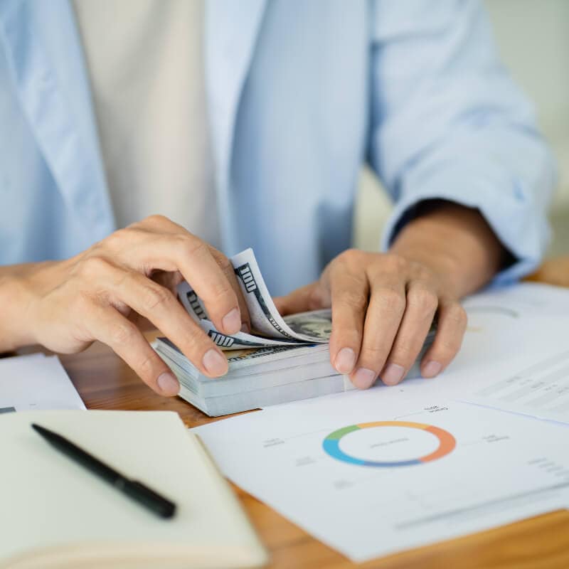 Person counting a stack of US dollar bills at a desk with financial documents, a chart, and a pen.