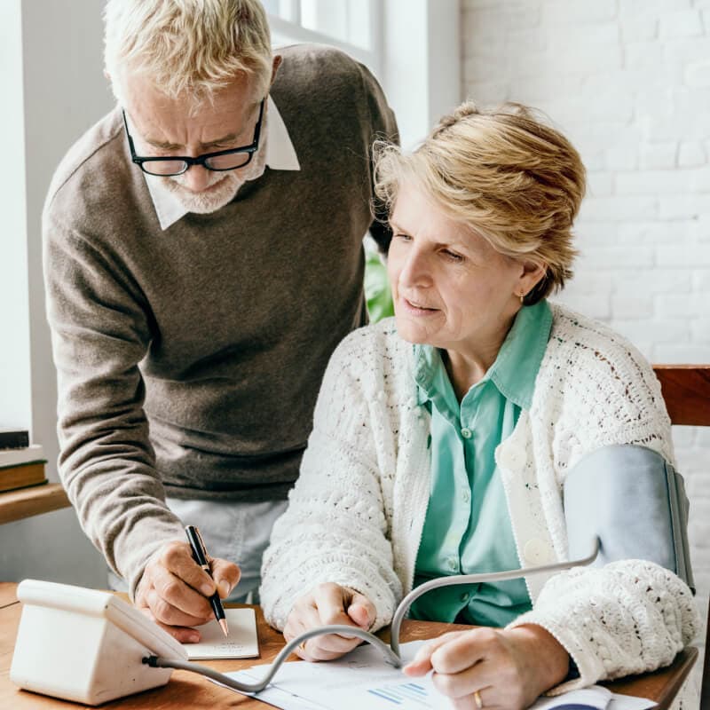 An older woman checks her blood pressure with a monitor while a man stands beside her and writes on a notepad.