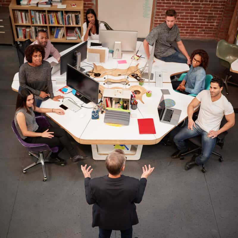 A group of people sit around large desks with computers in an office, listening to a person standing at the front, viewed from above.
