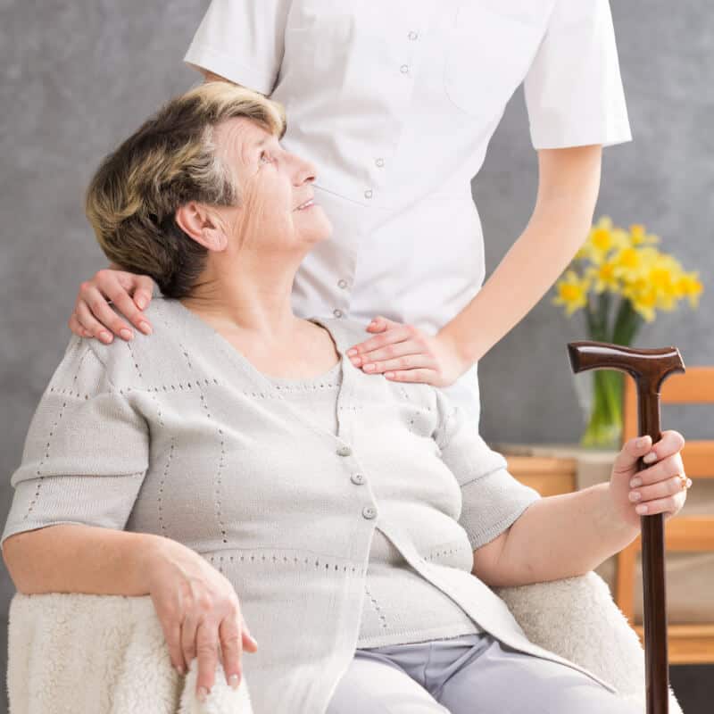 An elderly woman with a cane sits in a chair while a nurse in white uniform stands beside her, resting a supportive hand on her shoulder.