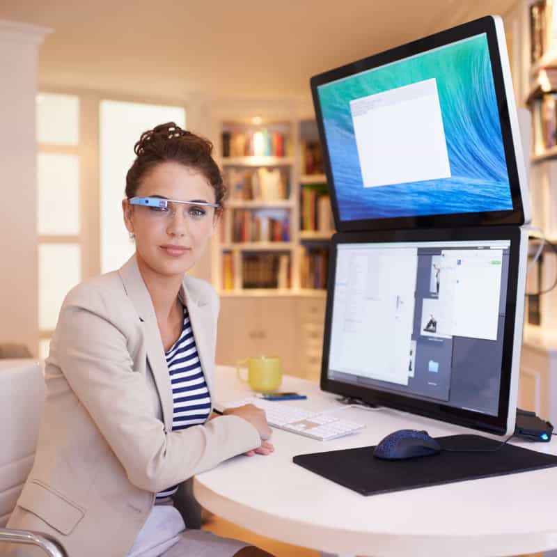 Woman wearing smart glasses sits at a desk with two large computer monitors, a keyboard, mouse, and a yellow coffee mug in a modern home office.
