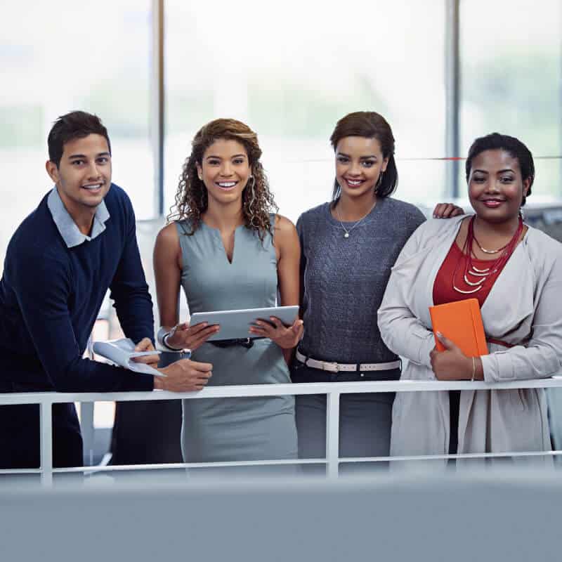 Four professionals stand together indoors, smiling at the camera. Two are holding a tablet and a notebook. They are dressed in business attire in a modern office setting.