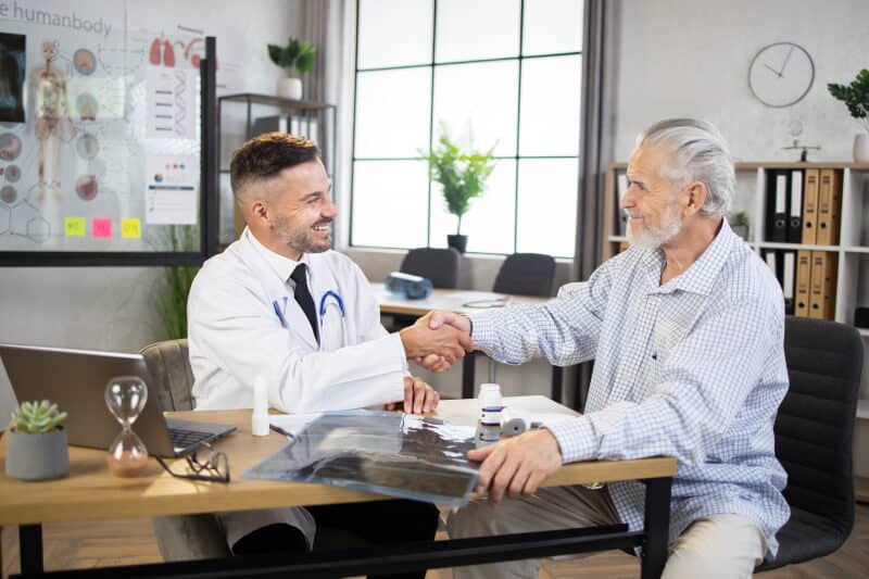 A doctor and an older male patient are sitting at a desk in a medical office, smiling and shaking hands during a consultation.