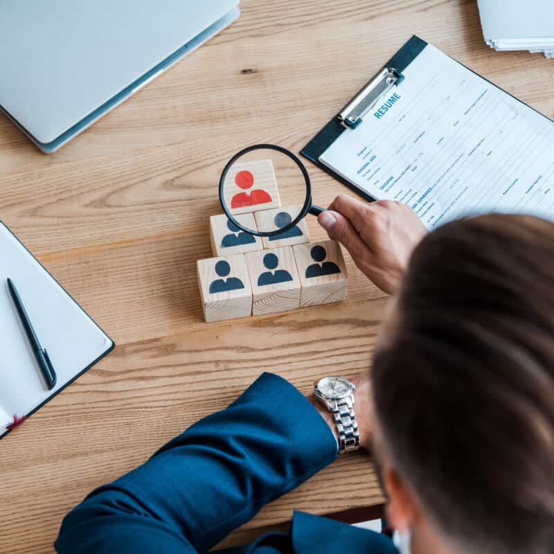 A person examines wooden blocks with human icons using a magnifying glass, focusing on a red figure, with a resume and office items on the desk.
