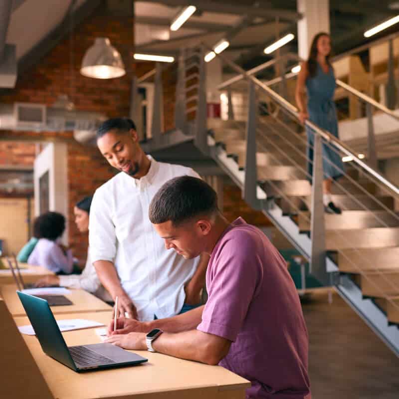 Two men collaborate at a high table with a laptop, while others work and a woman descends a staircase in a modern, open office space.