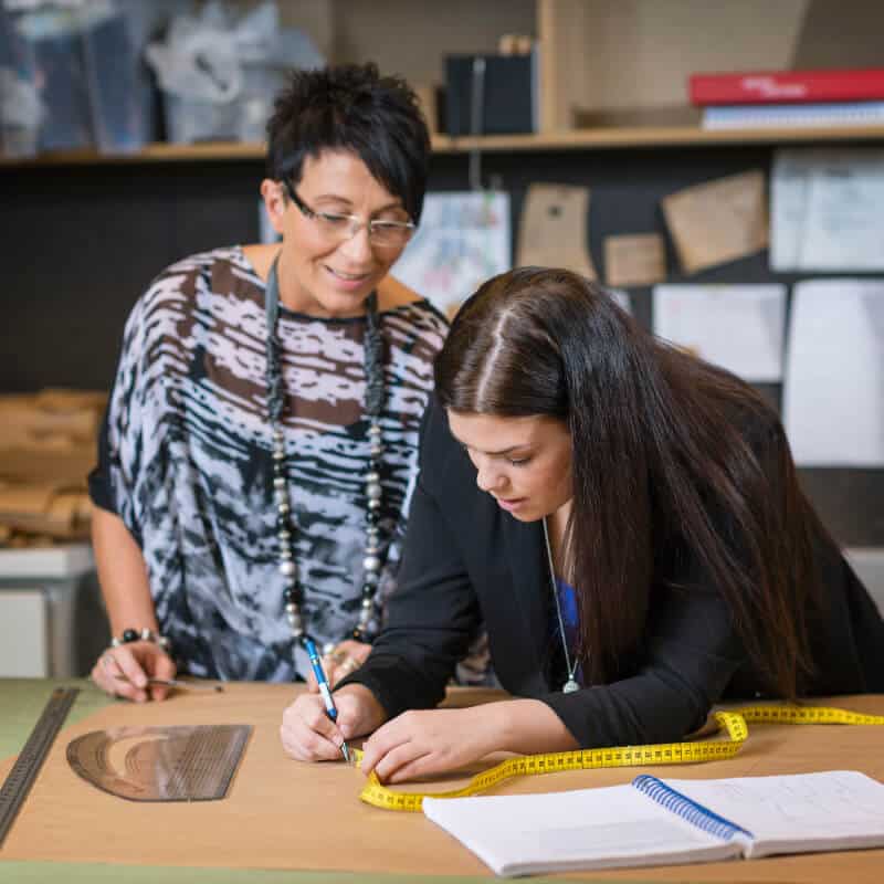 Two women work at a table; one writes on paper next to a measuring tape and curved ruler, while the other stands beside her, observing. Shelves and papers are visible in the background.