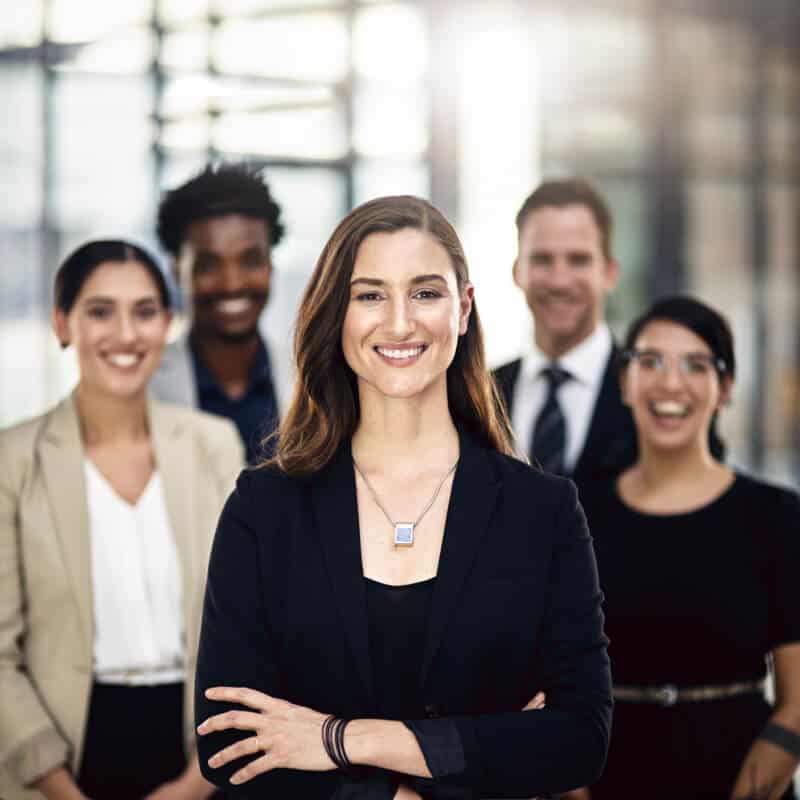 A confident woman in business attire stands in front of four colleagues, all smiling, in a modern office setting.