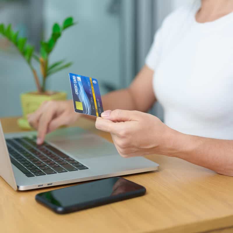 A person uses a laptop while holding a credit card, with a smartphone and a small potted plant on the wooden table.