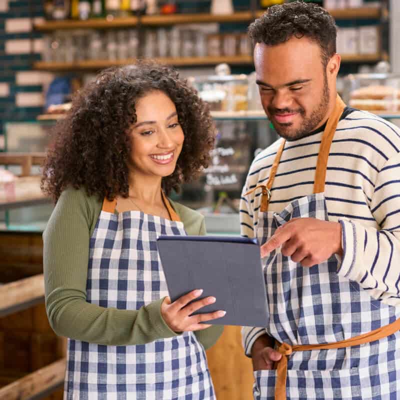 Two people wearing aprons stand in a café, looking and smiling at a tablet together. Shelves with jars and cups are visible in the background.
