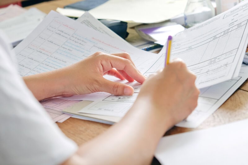 Person holding documents and filling out forms with a pencil at a desk cluttered with papers.