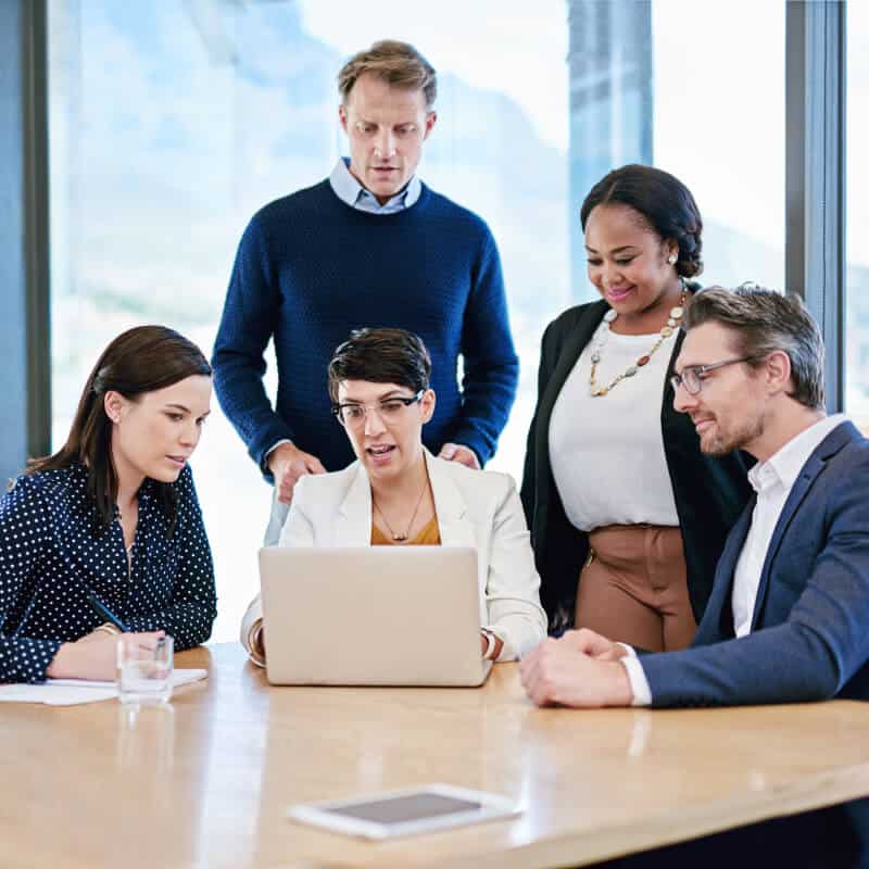 Five people gather around a table, looking at a laptop screen during a meeting in a modern office with large windows.