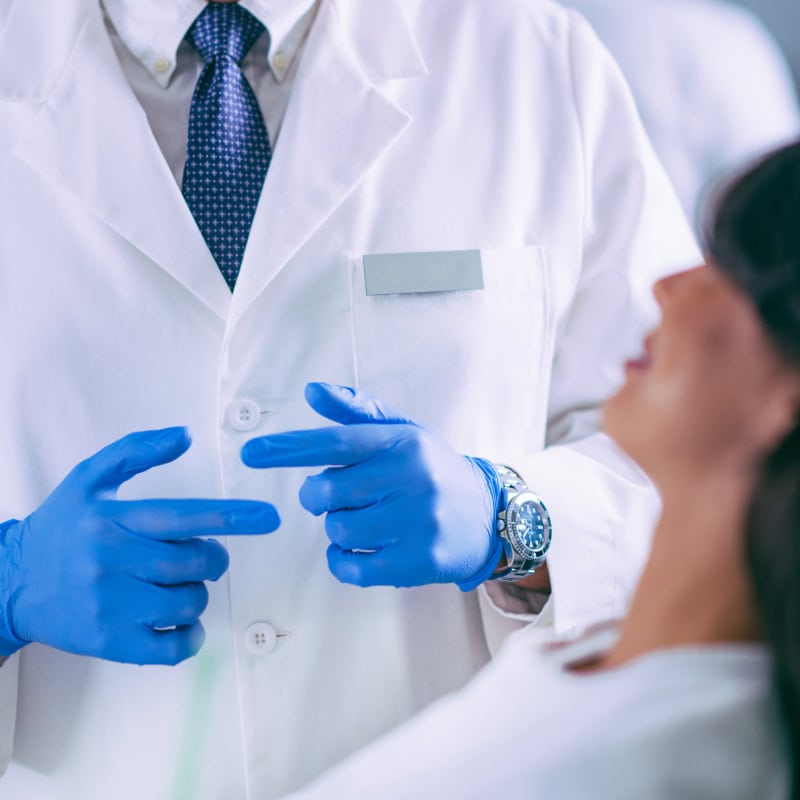 A healthcare professional in a white coat and blue gloves talks to a seated patient, with focus on the provider’s hands and upper torso.